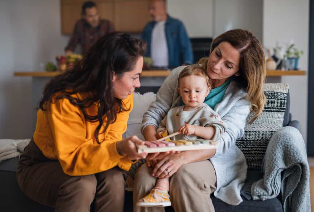 Two women sit with a toddler on a couch, one holding a book. In the background, two men discuss SNAP and SSI benefits in the kitchen.