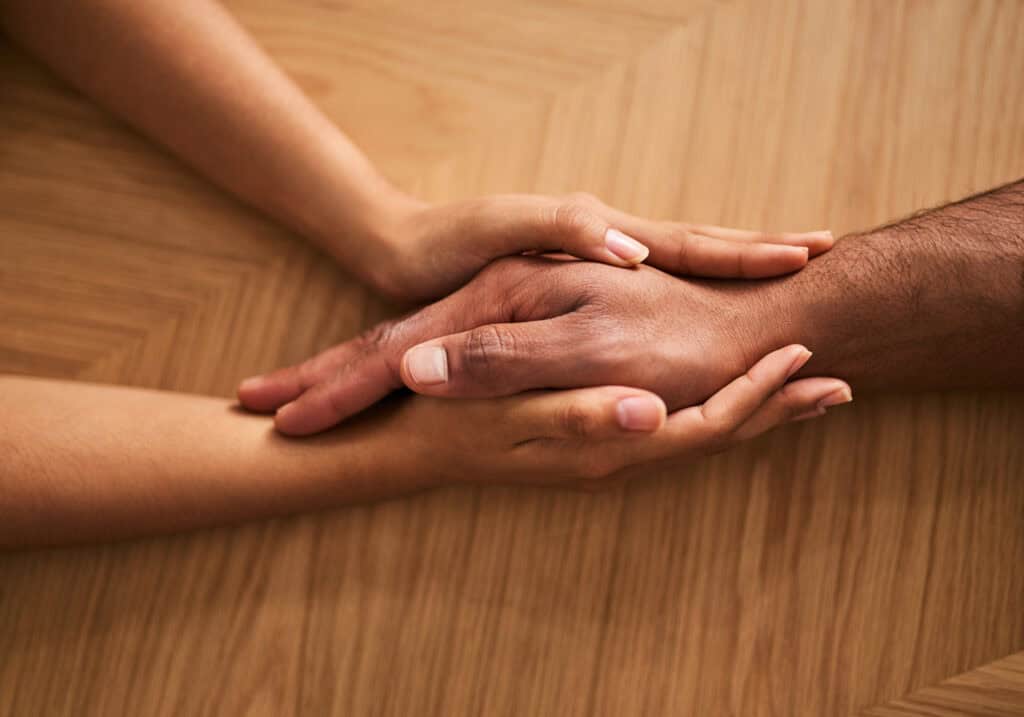 Two hands, one light-skinned and one dark-skinned, clasped together in support, rest on a wooden surface, symbolizing unity and shared strength in navigating life’s challenges like SNAP and SSI benefits.